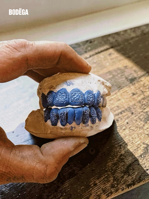 A close-up of a hand holding a dental mold or model with blue painted teeth on a rustic wooden surface. The brand name BODËGA appears in the top left corner.